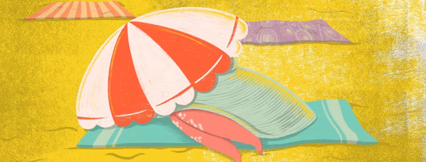 A person hiding under a beach umbrella and a comically large floppy summer hat while laying on a beach towel.
