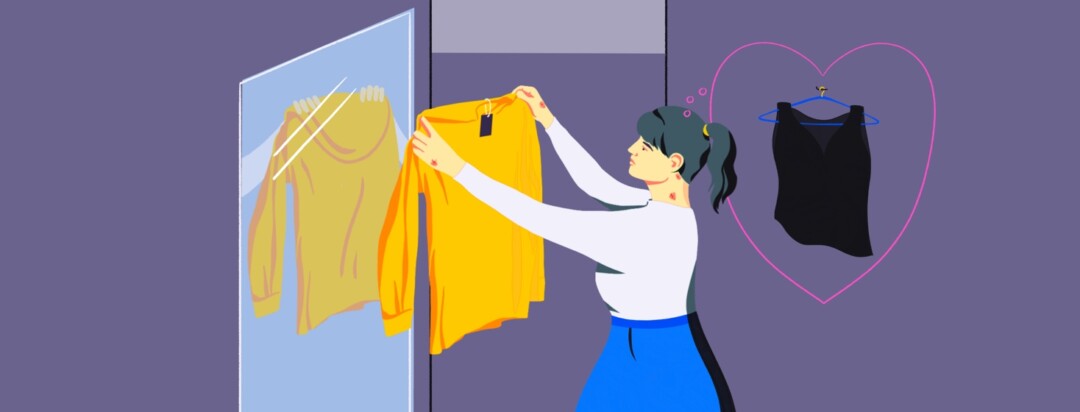 A woman stands in a changing room holding up a yellow blouse, a black blouse is on a hanger behind her with a heart around it